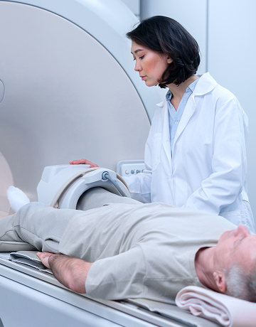 A patient is lying down and waiting for a CT scan of their knee while a radiology technician stands by the sliding table