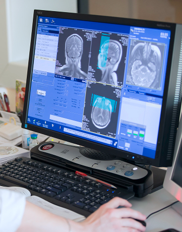 A radiologist sits at a desk and reviews MRI scans on their computer
