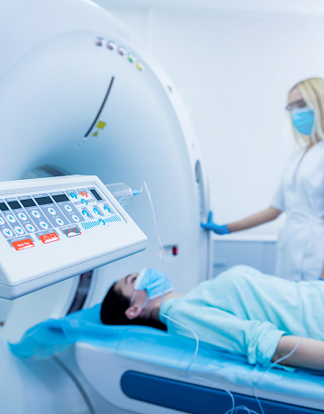 A patient lies on a table at a PET machine while a radiology technician pushes buttons to prepare the diagnostic imaging exam