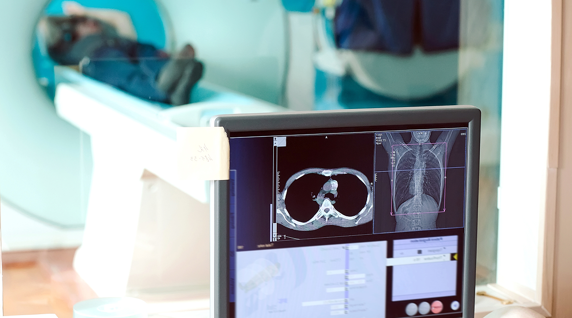 A computer screen shows the live results of an MRI scan while a patient moves through an MRI machine in the next room