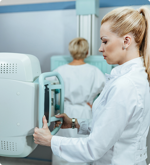 A physician holds and controls an X-ray machine while a patient stands in the background getting scanned