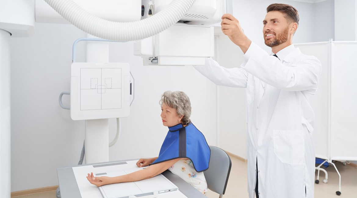 A patient sits and rests her arm on a table while a radiology technician prepares the machine for the diagnostic imaging exam