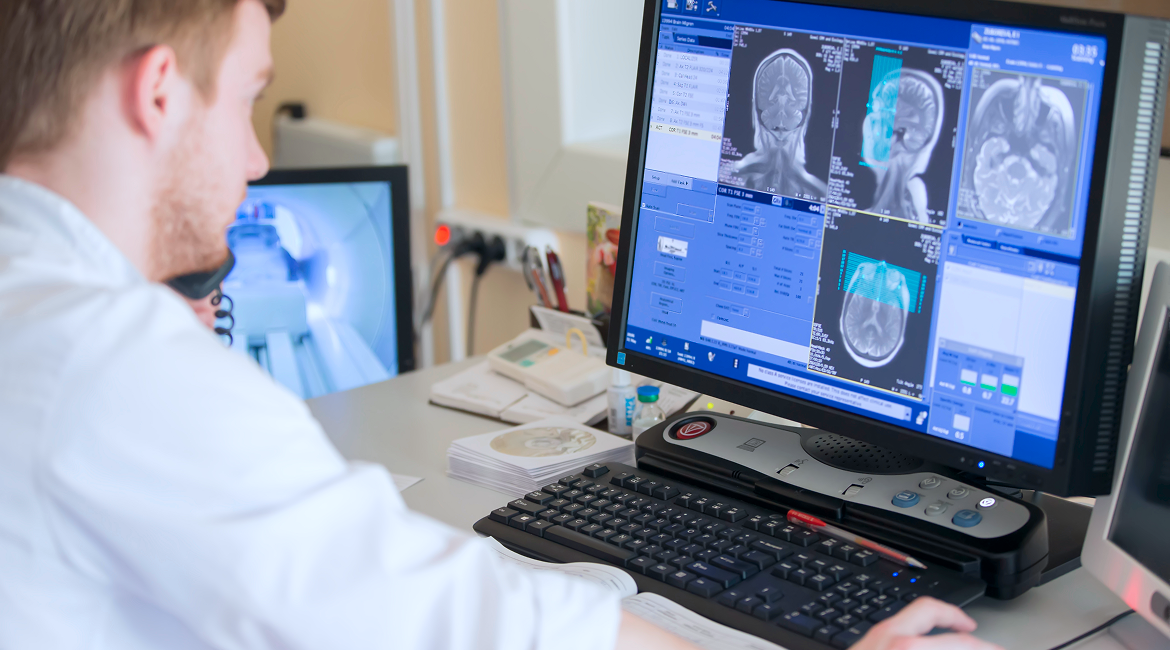 A radiologist sits at a desk and reviews nuclear medicine scans on their computer
