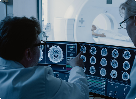 Two radiologists watch the live results of a CT scan on a screen while a patient moves through a CT machine in the next room