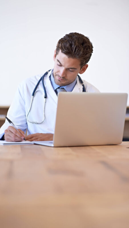 A physician has their laptop open while they sit at a table and take notes