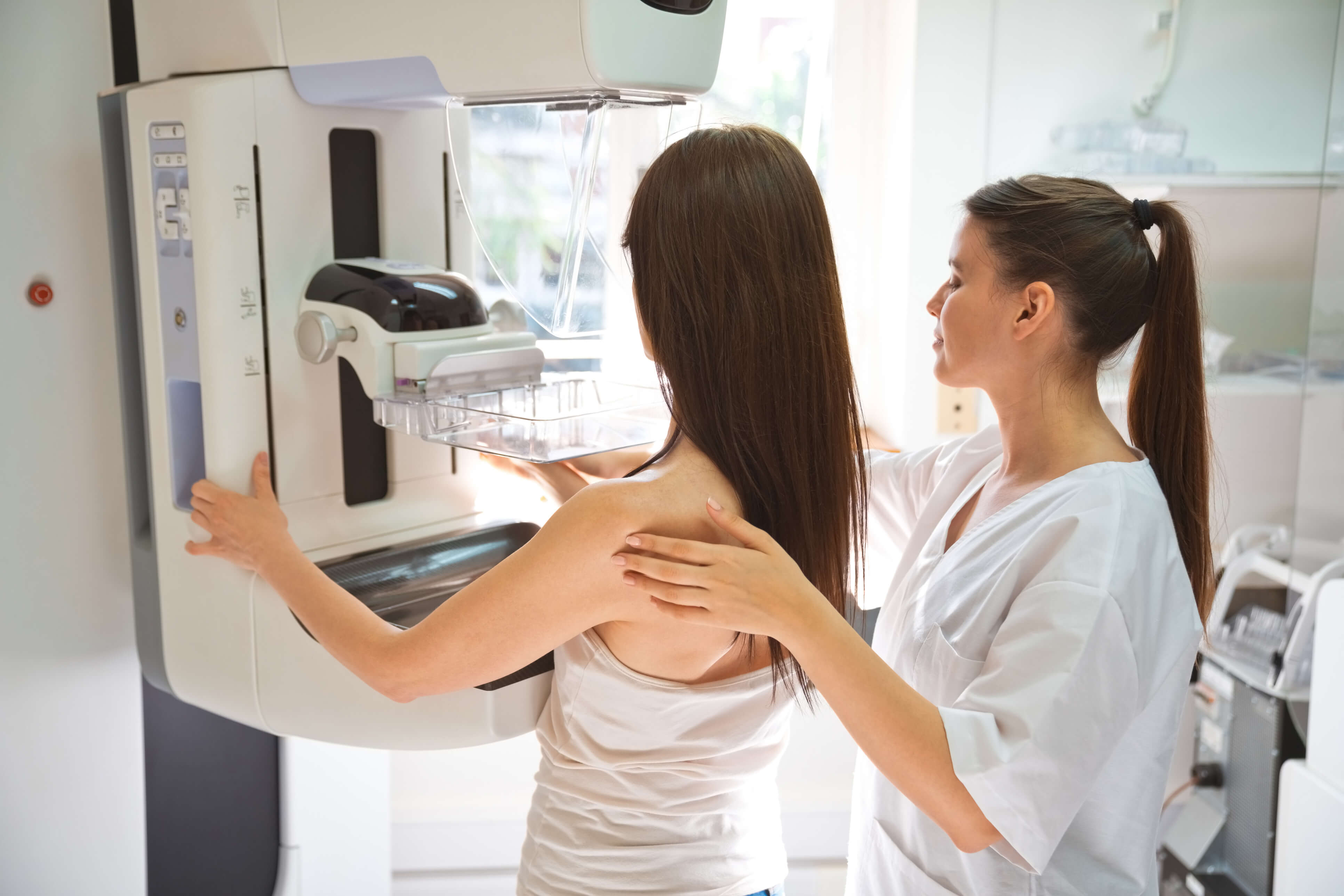 A female patient stands at a mammography machine while a radiology technician holds her shoulders and adjusts her position